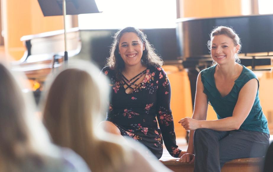 A music teacher and student sit on a stage in front of a grand piano while smiling to other students in the audience.